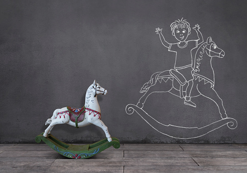 Side view of wooden rocking horse on wooden floor and little boy playing with rocking horse sketched (chalk drawing) on the wall.