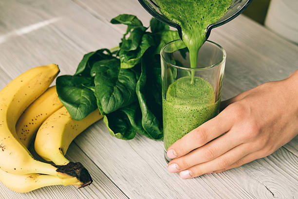 Female hand pours a smoothie of banana and spinach Female hand pours a smoothie of banana and spinach in glass on a wooden table spinach stock pictures, royalty-free photos & images