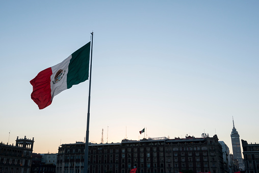 Mexico City, Mexico - October 28, 2014: A large Mexican flag flies at sunset in Mexico City's main plaza called the Zócalo. The highrise in the bottom right is the Torre Latinoamericana.