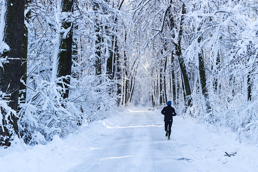 Man dressed in black sport equipment with blue jogging shoes running in the forest in winter time on a path covered by heavy snow. Horizontal scene with a forest covered by snow.