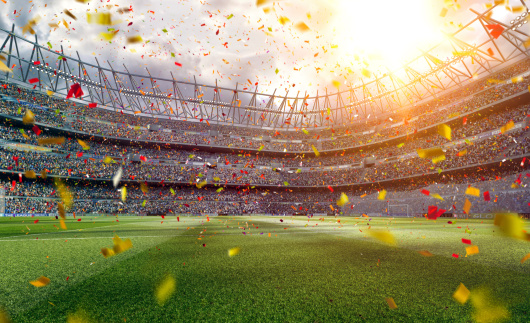 Panoramic view of soccer stadium with crowd on a sunny day.  The soccer field in the lower portion of the image has white lines running vertically and horizontally.  Portions of its green grass are shaded, while other parts are exposed to sunlight.  Stadium seating stretches across the middle portion of the image, and the seats are filled with spectators.  Bits of red and yellow confetti paper rain down from the sky and onto the field.  The sun is a large ball of glowing bright yellow above the upper right corner of the stadium.  Storm clouds billow above the stadium, appearing darkest in the upper left section.