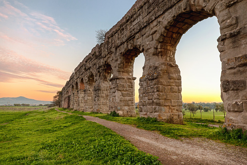 Arches of an ancient Roman aqueduct, made of blocks of tufa. A path runs along the property in a green park in the outskirts of Rome.