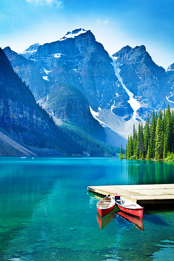 Lake Moraine in the Banff National Park of Canada, with its emerald water and mountain range of the Canadian Rockies. Red rental canoes moored against the dock with the dramatic natural scenic mountain lake in the background. Photographed in vertical format.