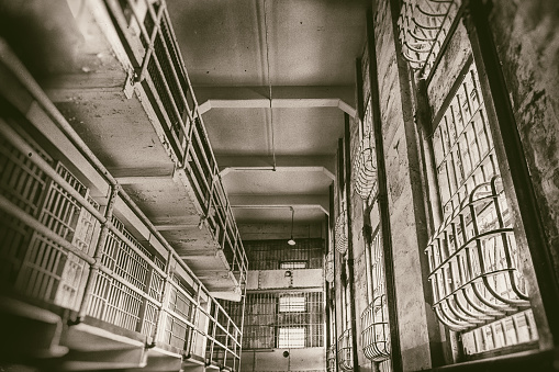 Perspective photo of a row of old prison cells with natural light spilling in from above; cell block at Alcatraz.  Color toning and texture added for vintage effect.