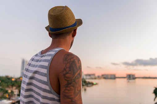 This is a horizontal, color photograph of a young Brazilian man in his 20s standing outside. He looks over the water of the Intracoastal as the sunset begins to change the colors of the sky. Photographed from a rearview. He dresses in a tank top. A tattoo on his arm shows.