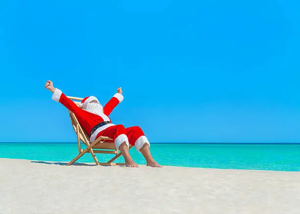 Christmas Santa Claus hands up enjoy sun on deck chair at ocean sandy tropical beach - New Year vacation and travel destinations in hot countries concept