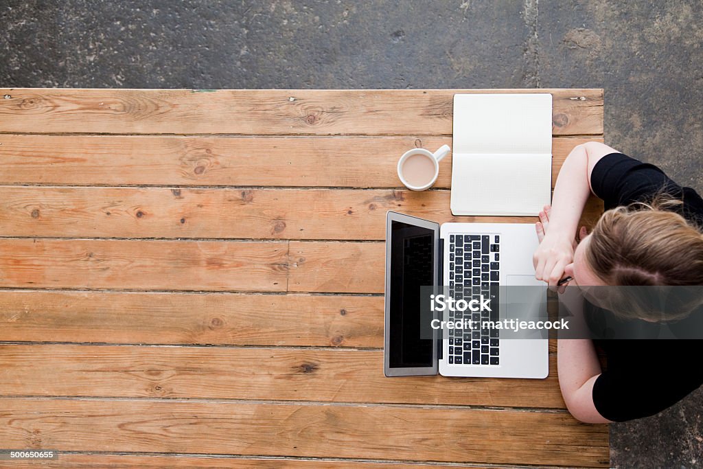 Woman working at a laptop photographed from directly above A woman is working at a rustic wooden desk, in front of her is a laptop, note pad and cup of tea. It has been photographed from directly above with the woman and objects on the right hand side of the image and blank space on the left hand side. Above Stock Photo