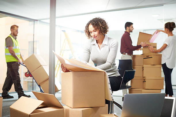 new office move day a young female entrepreneur sits at her laptop in an empty office surrounded by moving in boxes but as yet no furniture. Behind her colleagues are organising files, and a male co-worker is chatting to a contractor as to where the furniture needs to be going  . She is looking through the layout of the office on some plans . company relocation stock pictures, royalty-free photos & images