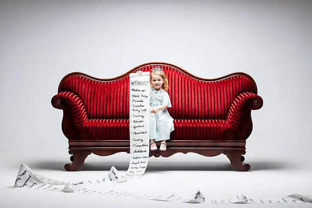 3 year princess girl sitting on red vintage sofa and holding an extraordinary long list of material whishes.