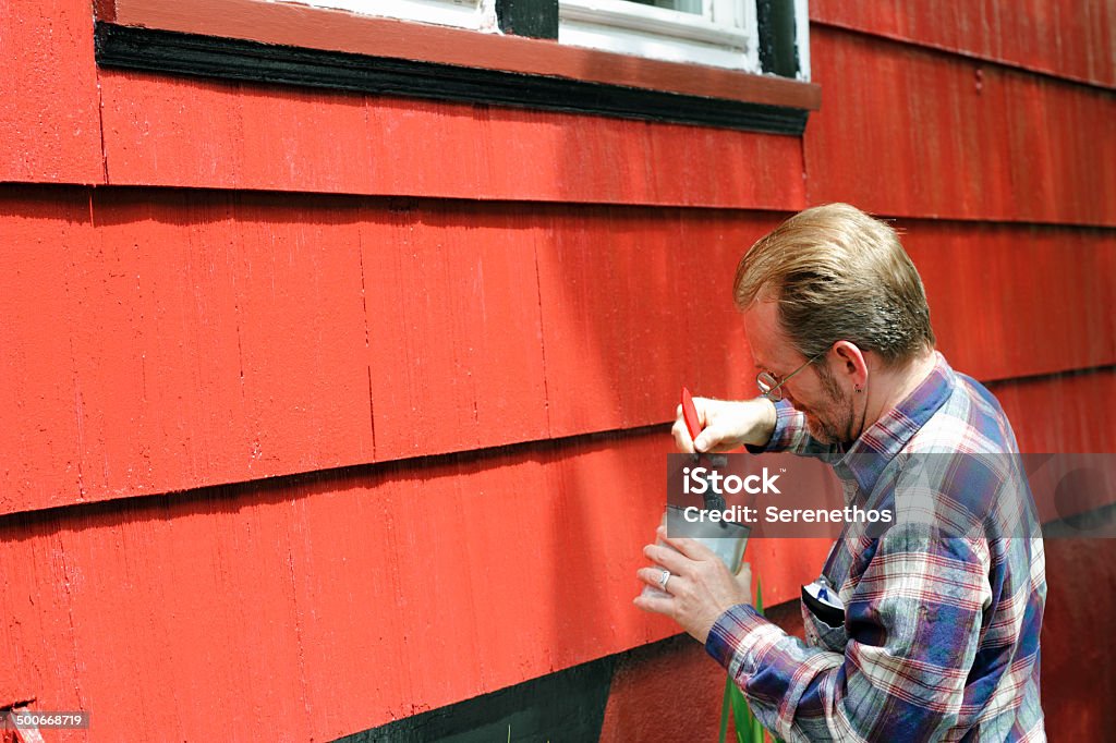 Home Improvement Painting Mature male dipping a paintbrush in a can of black paint in order to touch up trim around the windows of his house on a sunny day.. Painting - Activity Stock Photo