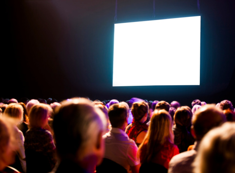 Crowd stares at a large screen in a dimly lit room.  The crowd is sitting in organized rows, and the screen is shining so brightly that it appears white.