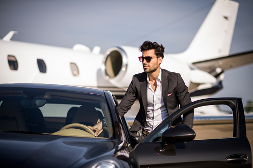 Young well dressed man with sunglasses, looking like famous person or other celebrity, exiting from the black car after arriving at the airport track. Private jet airplane is in the background.