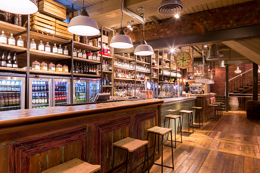 Interior of a traditional British food and drinks establishment with a modern twist. No people. Bar stools and bar counter seen with overhead lighting. Rustic look with beer bottles behind the bar.