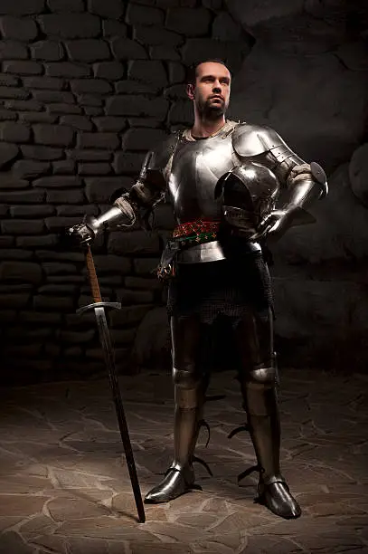 Medieval Knight posing with sword  and helmet in a dark stone background. Full-length portrait.