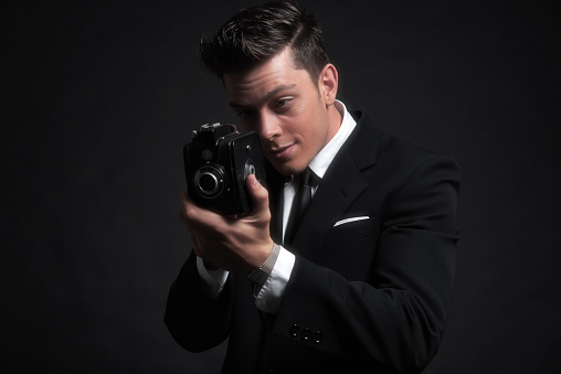 Retro fifties male photographer with vintage camera. Wearing black suit and tie. Studio shot.
