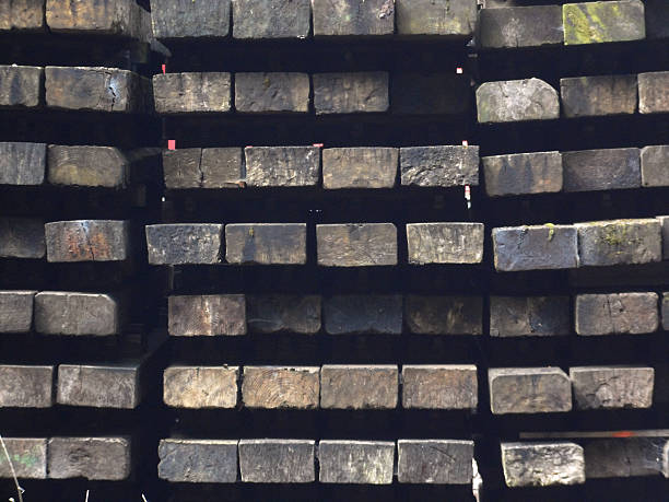 Large pile of weathered timber railroad sleepers stacked up beside a railway line.