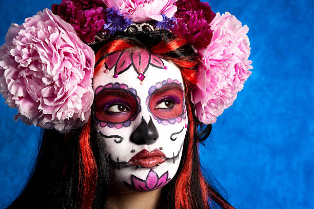 Horizontal studio shot on blue of young woman in red and pink sugarskull Day of the Dead makeup, closeup looking away.