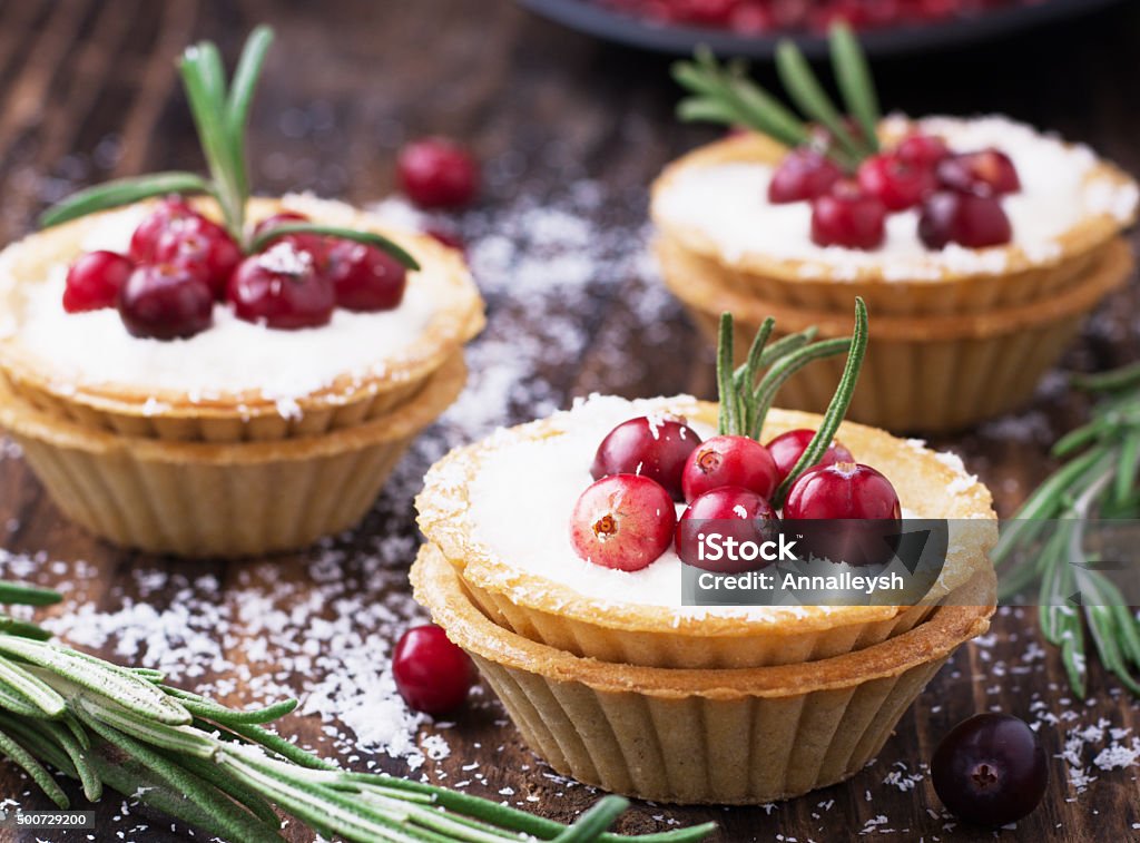 Tartlets of pastry with cream and fresh berries ripe cranberries Tartlets of pastry with cream and fresh berries ripe cranberries and rosemary leaves sprinkled with coconut on the texture wooden background. selective Focus Dessert - Sweet Food Stock Photo