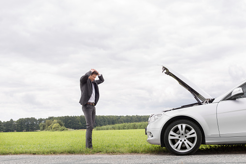 Sad young businessman standing with hands on head near broken car at countryside