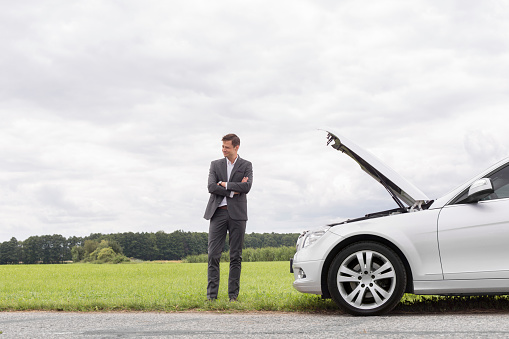 Young businessman standing arms crossed by broken down car at countryside