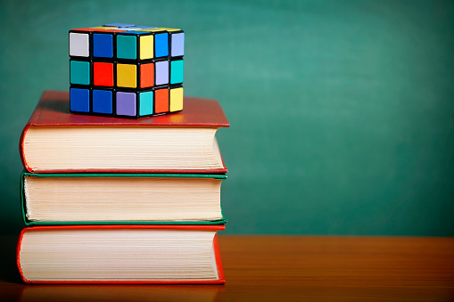 Ulyanovsk, Russia - November 30, 2015: Stack of books with a Rubik's cube on a wooden desk. Green chalkboard background. Shallow depth of field. Space for copy. Can illustrate the concept of education, logic, learning, etc.