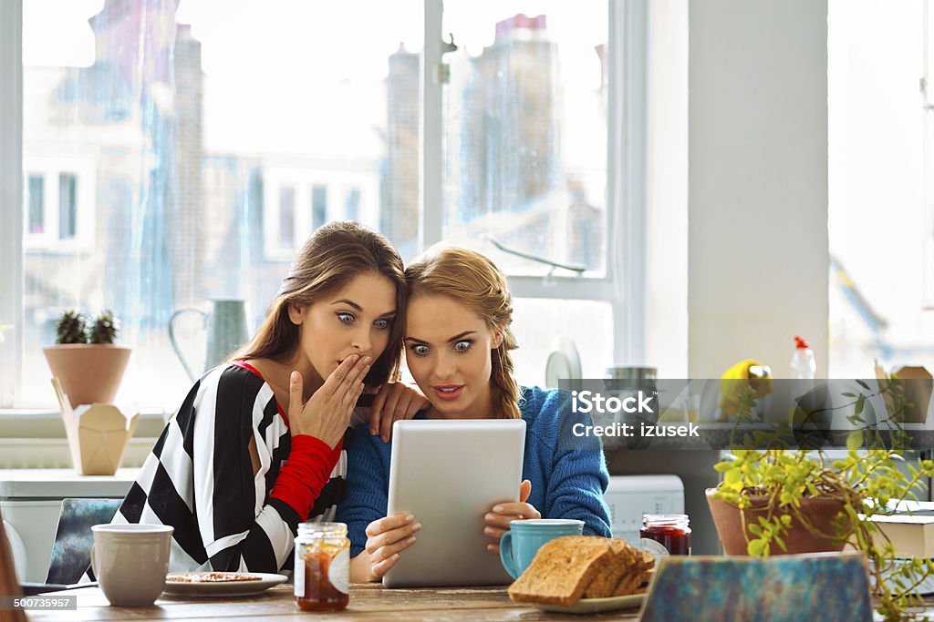 Girls using digital tablet Two friends sitting at the kitchen table and using digital tablet together.  Gossip Stock Photo