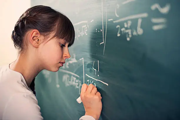 Little mathematics genius girl writing difficult mathematics equations on a green chalkboard. The gilr is smiling focused on solving the equation.
