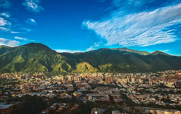 Photograph of Caracas, a Venezuelan city under siege from organized crime and corruption, shot during a beautiful sunny November afternoon displaying the vibrant contrast between the inherent beauty of the city (as shown from a far wide angle shot) and the internal dangers of its daily life.
