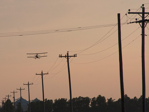 A bi-plane flies over power lines at sunset.