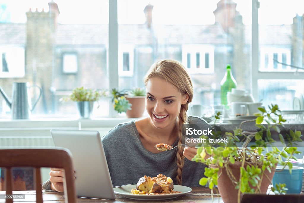 Young woman at home Young woman sitting at the table in the kitchen at home, eating a jacket potato with beans and cheese and using a digital tablet. Baked Potato Stock Photo