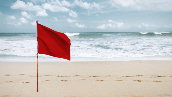 The Australian and Australian Aboriginal flags on flagpoles with the background of Bondi Beach, Sydney.  A woman is walking along the beach.  This image was taken on a partly cloudy afternoon at low tide on 31 August 2023.