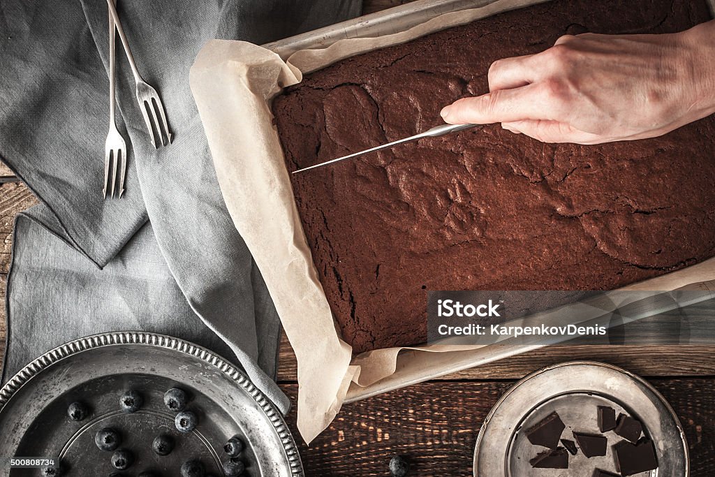 Cutting chocolate brownie horizontal Chocolate brownie on the baking tray  with blueberry , knife in the hand , chocolate , forks and  old vintage plates  on the wooden table horizontal 2015 Stock Photo