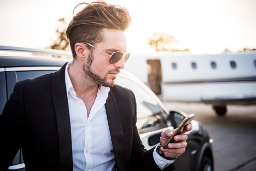Young well dressed man with sunglasses holding his smart phone. He looks like a famous musician or other celebrity. Private jet airplane is in the background as well as bright sunlight.