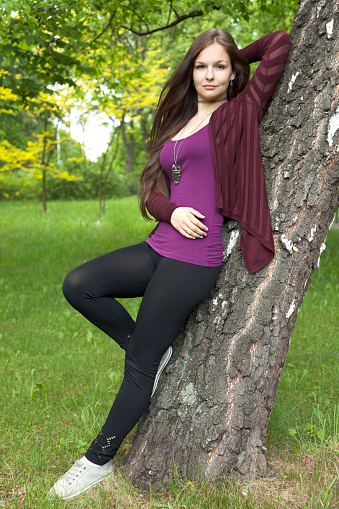Portrait of young woman in nature. She is leaning on birch and looking into the camera.