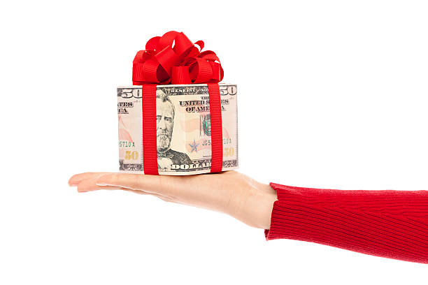 Hand Offering Cash Money Gift of U.S. Currency on White Subject: Horizontal view of a human hand holding a square gift box wrapped with paper of a U.S. one hundred dollar bill, tied with red ribbon and a fancy bow. The bow color suggests a Christmas gift, bonus, birthday present, or special occasion. Isolated on a white background. gift card $100 stock pictures, royalty-free photos & images