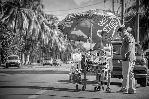 Havana, Cuba - November 6, 2015. Street scene from suburbs of Miramar, not far from old Havana. Older woman selling groceries out of her shopping cart. She is talking to a male customer standing next to her on the urban street. Huge umbrella is tucked in, protecting her from afternoon sun. Wide urban street with cars in the background.