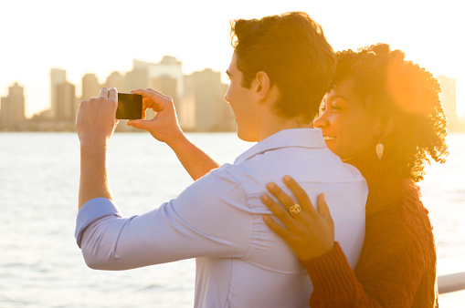 Romantic couple tourists taking photo and having fun. Young happy couple taking photo of the river and skyscrapers with smartphone. Happy young couple embracing and taking photo at river side during sunset.