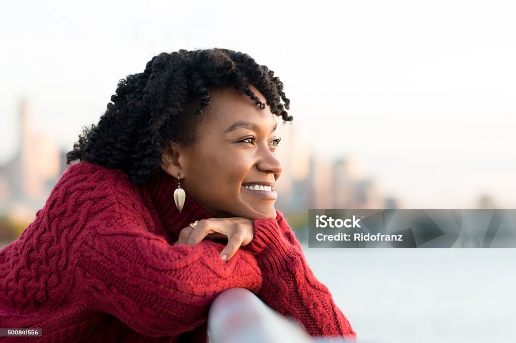 Woman thinking outdoor Close up portrait of a young happy african woman leaning on the banister of a bridge near river. Happy young african woman at river side thinking about the future. Smiling pensive girl looking across river at sunset. Women Stock Photo