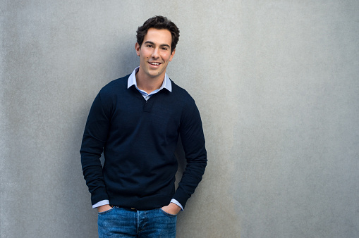 Happy man leaning on a grey wall. Handsome young man in casual leaning against a grey background and copy space. Young businessman looking at camera with hands in pocket.