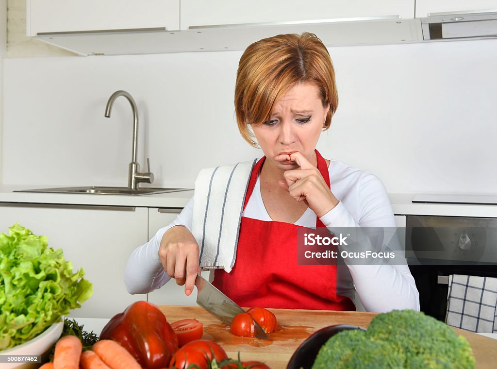 home cook woman suffering domestic accident  knife cutting finger young attractive home cook woman in red apron slicing tomato with kitchen knife  suffering domestic accident cutting and hurting her finger while cooking in pain face expression licking blood Table Knife Stock Photo