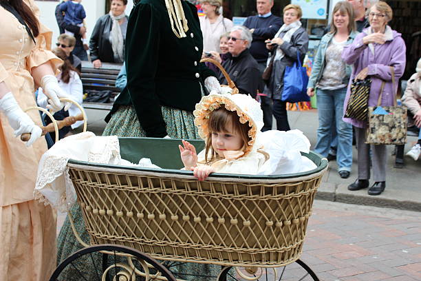 Mother and little daughter are proudly walking theirs babies in original vintage prams. Dressed in classic dresses in white and navy blue, they are in a public city park, in summer. Baby is 5 month, little girl is two. Trees in the background. Full length horizontal shot with copy space.