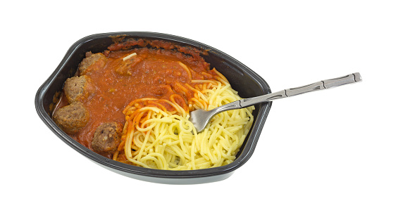 A microwaved spaghetti and meatball TV dinner in a black tray with a fork inserted into the pasta on a white background.