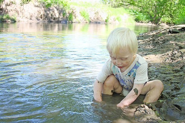 Young Child Sitting in the River During Summer a young boy child is sitting in on the beach of a river in the woods, playing in the mud on a summer day. people covered in mud stock pictures, royalty-free photos & images