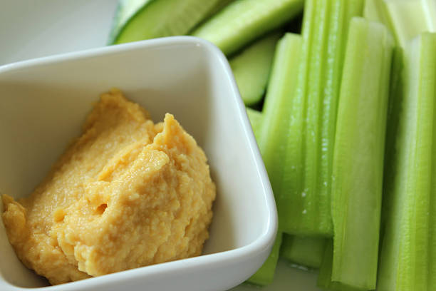 Photo showing a homemade hummus dip in a small square white bowl, served on a plate with some chopped celery and cucumber sticks (cut julienne style).  This healthy, low calorie snack is part of a low carb, healthy eating diet plan, eaten in between meals.