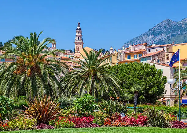 Catholic church's bell tower and colorful houses under blue sky as seen through green palms and trees in Menton, France.