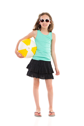 Young girl in teal shirt and black skirt posing with a beach ball under her arm. Full length studio shot isolated on white.