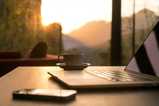 Nomad work Concept Image Computer Coffee Mug and Telephone large windows and sun rising, focus on coffee mug