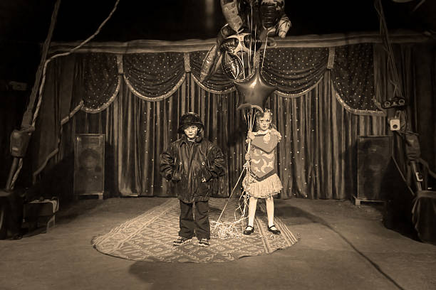 Young Boy Dressed as Clown Wearing Leather Jacket and Helmet Standing on Stage with Girl in Bright Costume Holding Strings of Foil Balloons, Monochrome Image Gives Old Time Feel