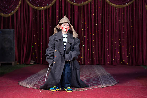 Talented boy standing and holding a sword while wearing oversized retro gray coat and Russian hat over modern casual clothes during theatrical play on stage