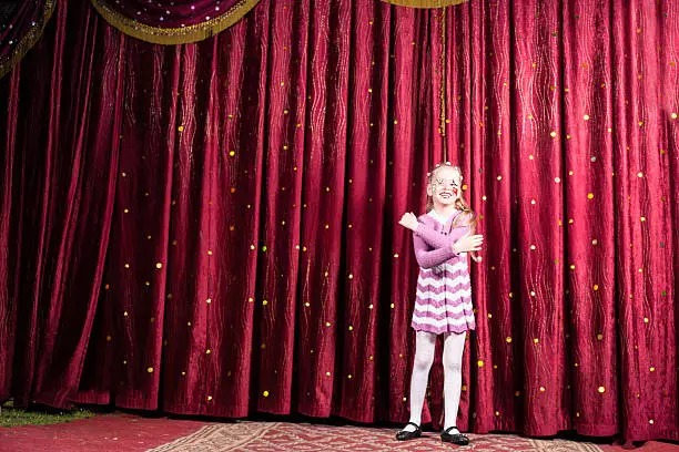 Full Length Portrait of Smiling Blond Girl Wearing Clown Make Up and Striped Dress Standing on Stage in front of Red Curtain